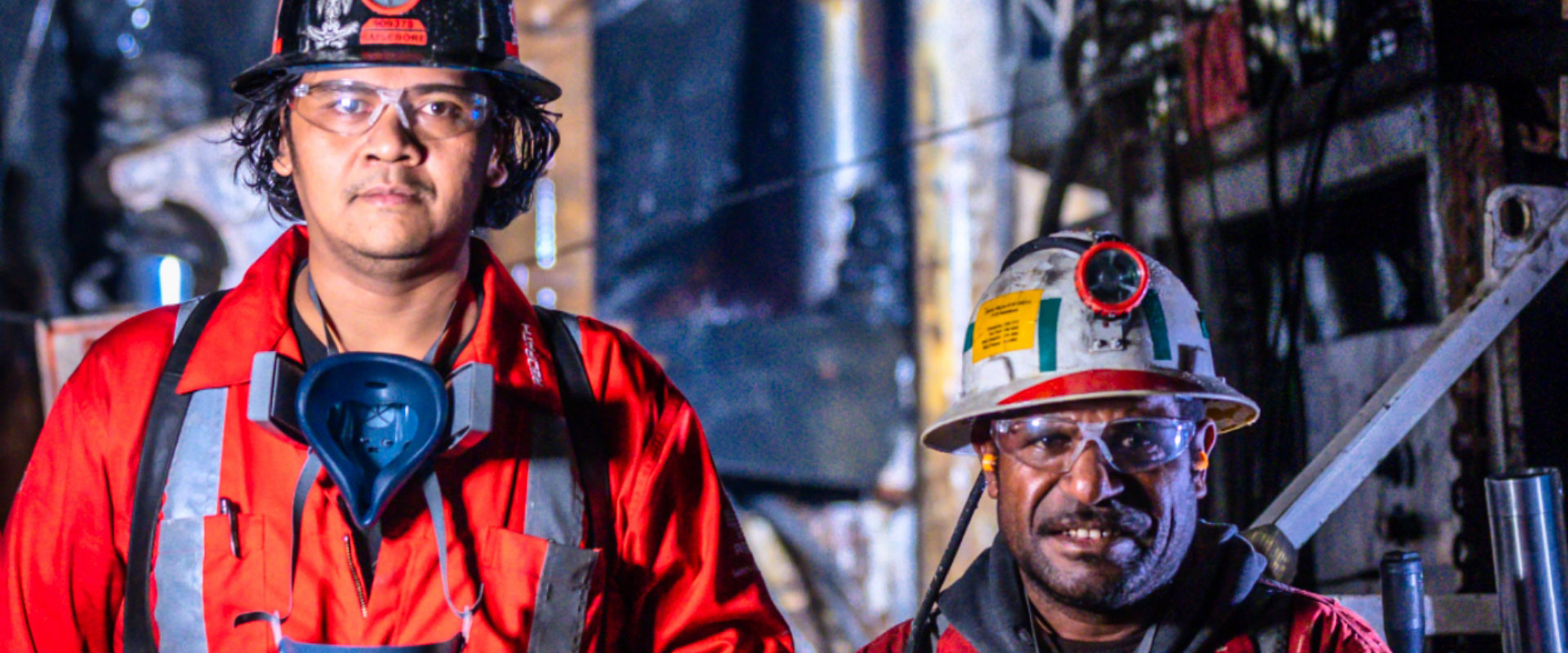 Dos mineros indígenas bajan por un pozo, vistiendo equipo de protección, mirando directamente a la cámara, transmitiendo una sensación de trabajo en equipo y dedicación dentro de un entorno de trabajo.